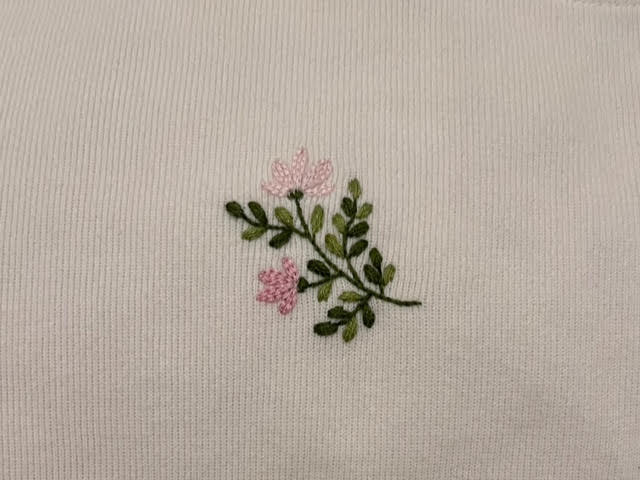 Photograph of embroidered fabric. The embroidery is green rounded leaves with two pink delicate-looking flowers.