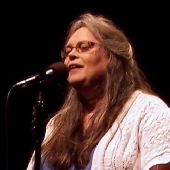 A person with long hair and glasses singing into a microphone

Description automatically generated with low confidence
